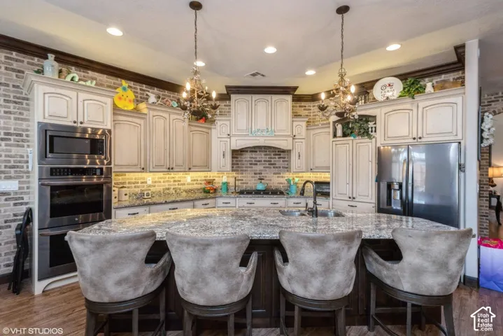 Kitchen featuring a large stone-surface island, cream-toned cabinetry with dark decorative molding, exposed brick walls, built-in stainless steel double ovens, and suspended decorative light fixtures