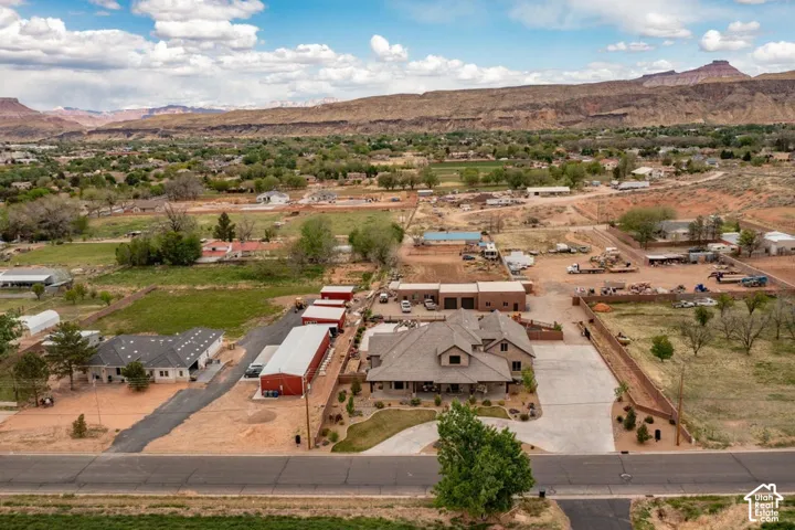 Aerial perspective of a multi-structure property featuring a main residence with a hipped roofline, multiple detached metal-roof workshops, extensive concrete driveway space, and a backdrop of mesa formations