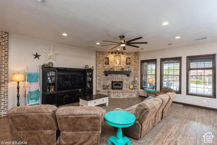 Corner fireplace with brick surround, wood-finish flooring, recessed lighting, ceiling fan, and large windows with dark trim