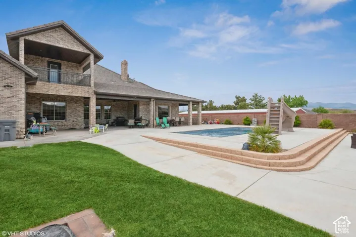 Brick exterior with a covered patio, concrete pool deck, in-ground pool with stone-finished steps, second-story balcony with metal railing, and a masonry perimeter wall
