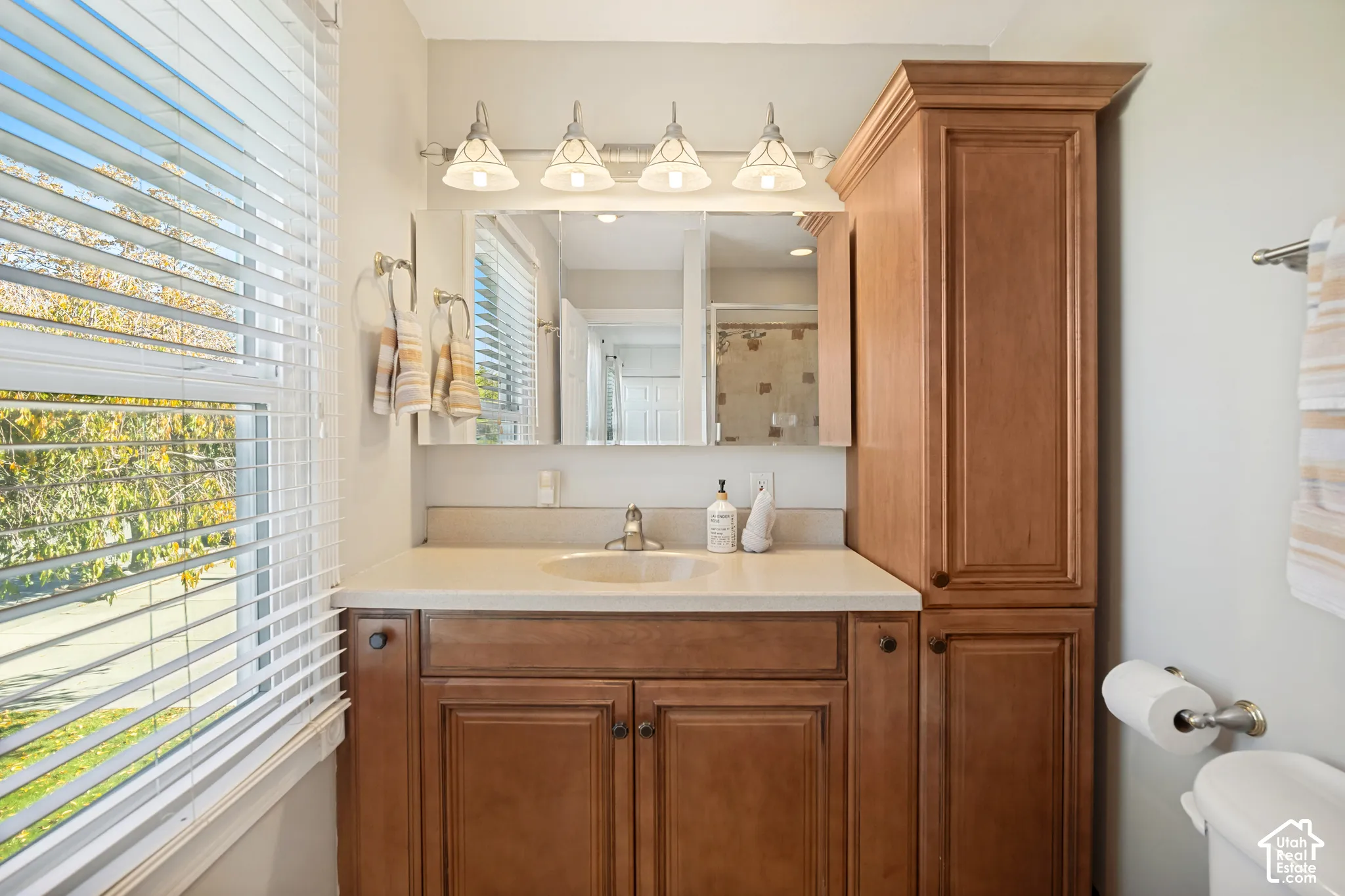 Bathroom vanity featuring wood-finish cabinetry, a tall built-in linen tower, a light-toned countertop with an integrated sink, and a four-light fixture