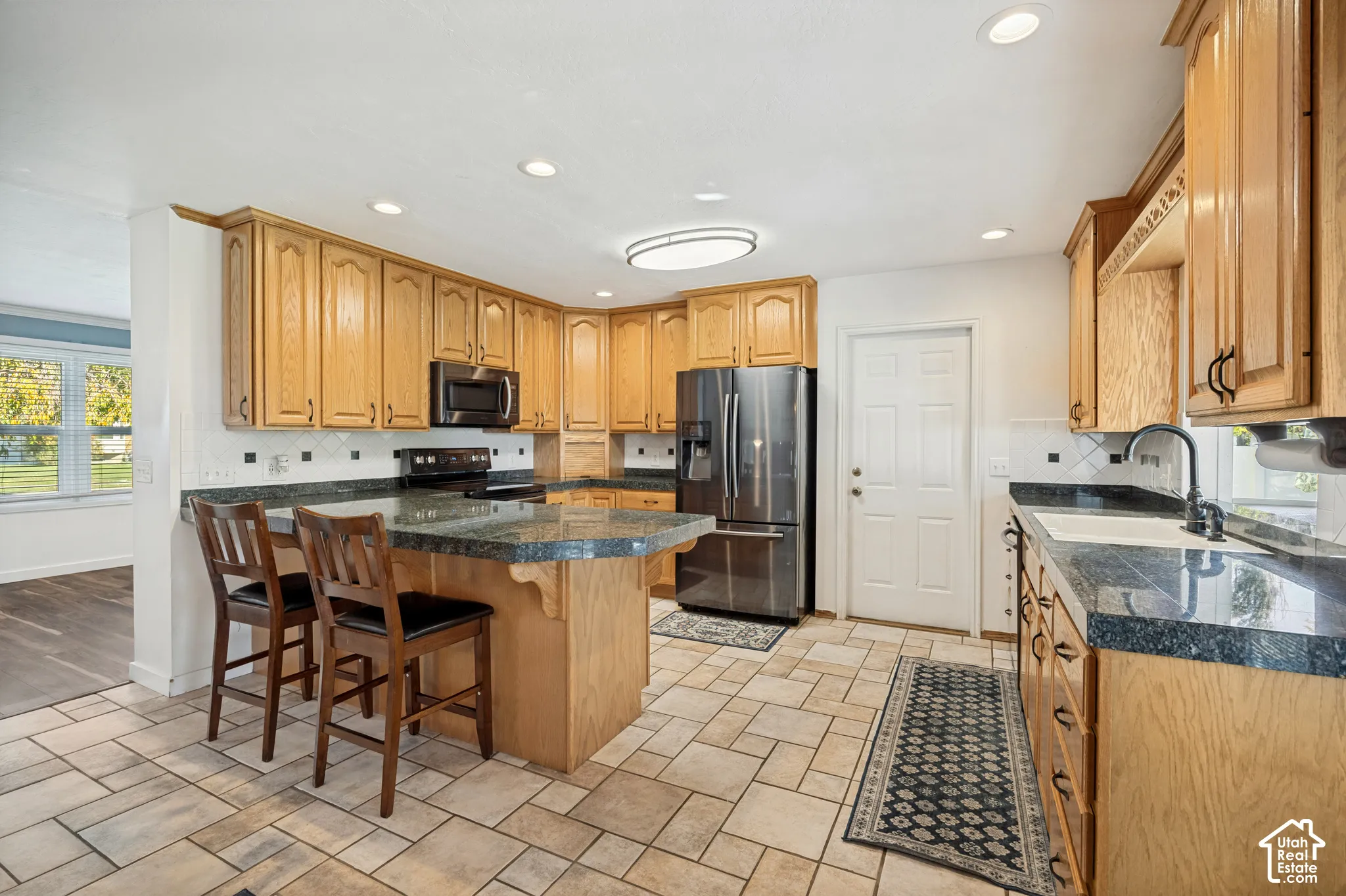 Kitchen featuring medium-tone wood cabinetry, stone-finish countertops, tile flooring, built-in stainless steel appliances, and a central peninsula with counter seating