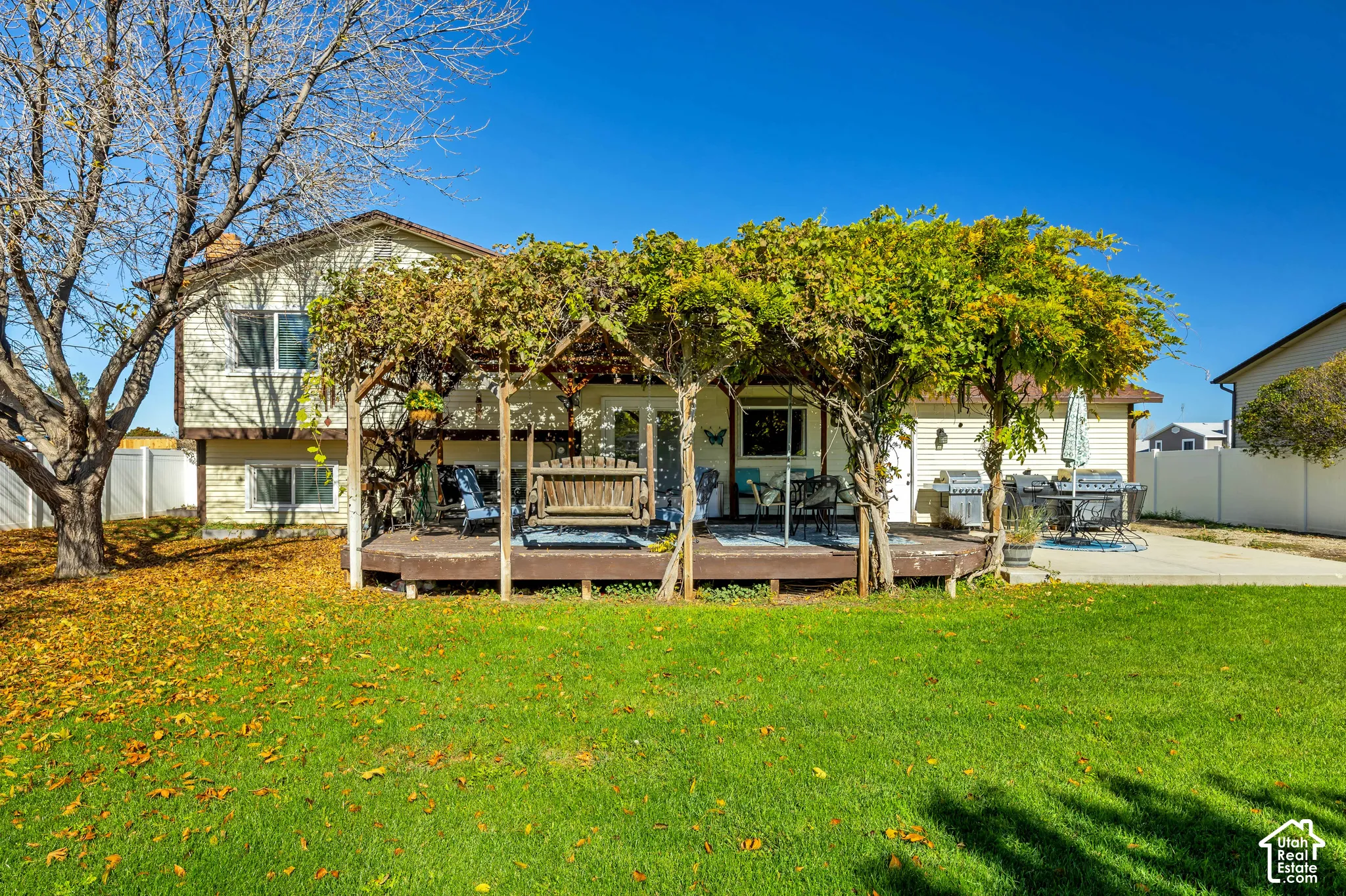Backyard featuring a raised wooden deck, a pergola covered in mature vines, a concrete patio, and white vinyl privacy fencing