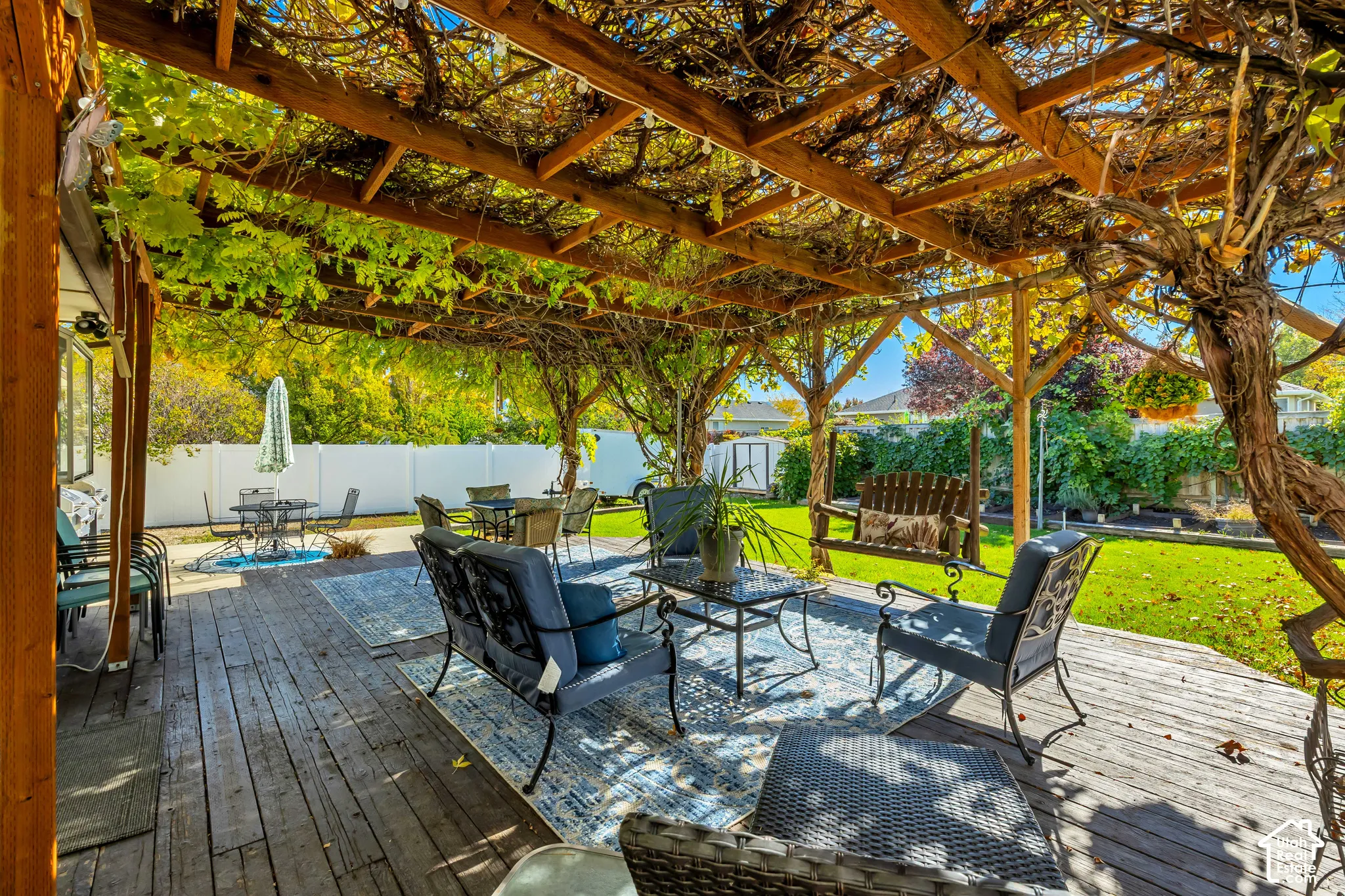 Spacious wooden deck featuring a natural wood-beam pergola with climbing vines, a manicured lawn, and a perimeter privacy fence