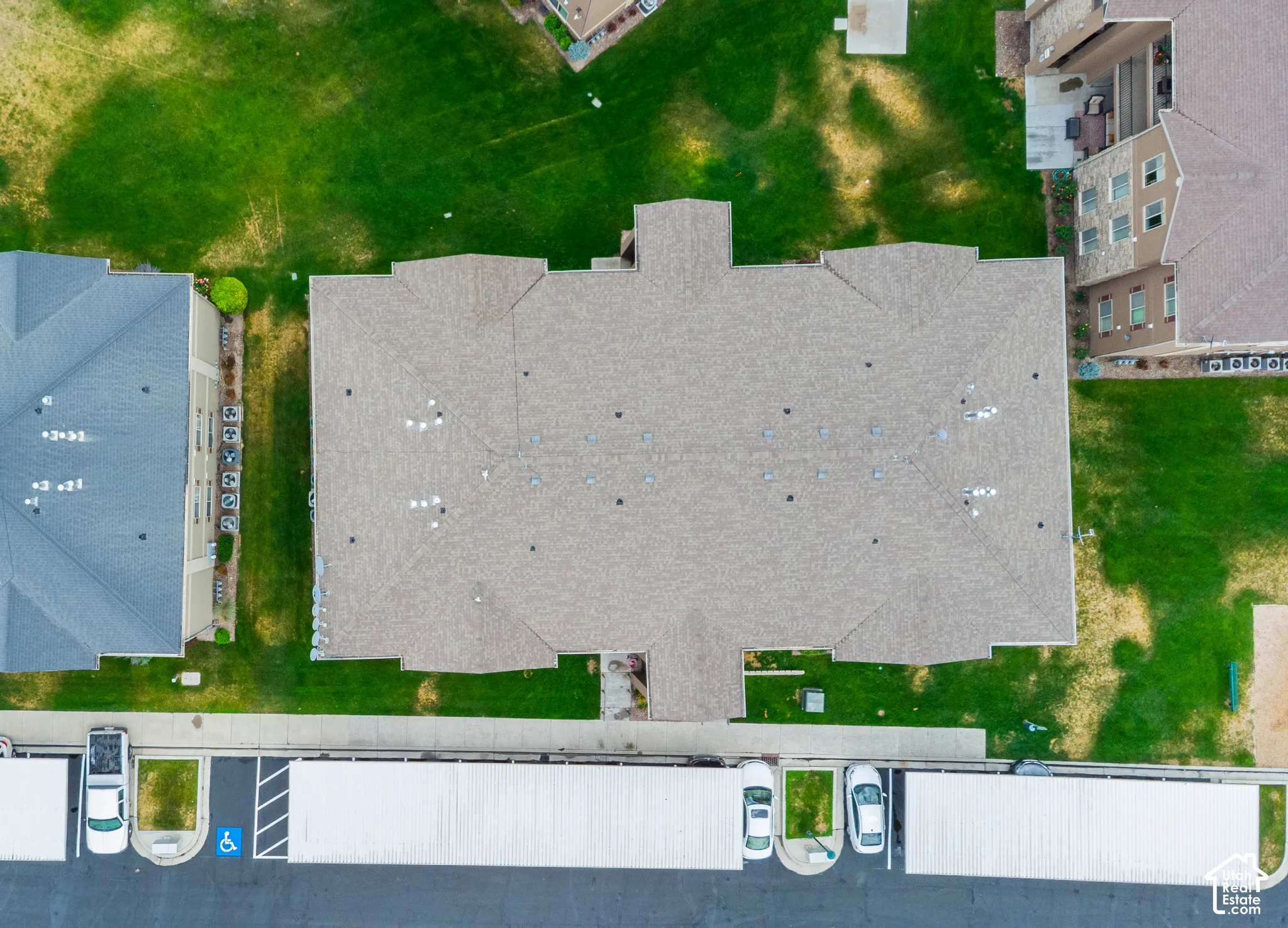 Aerial view of multi-unit building featuring a composite shingle roof, expansive surrounding lawn, paved parking lot, and covered parking structures