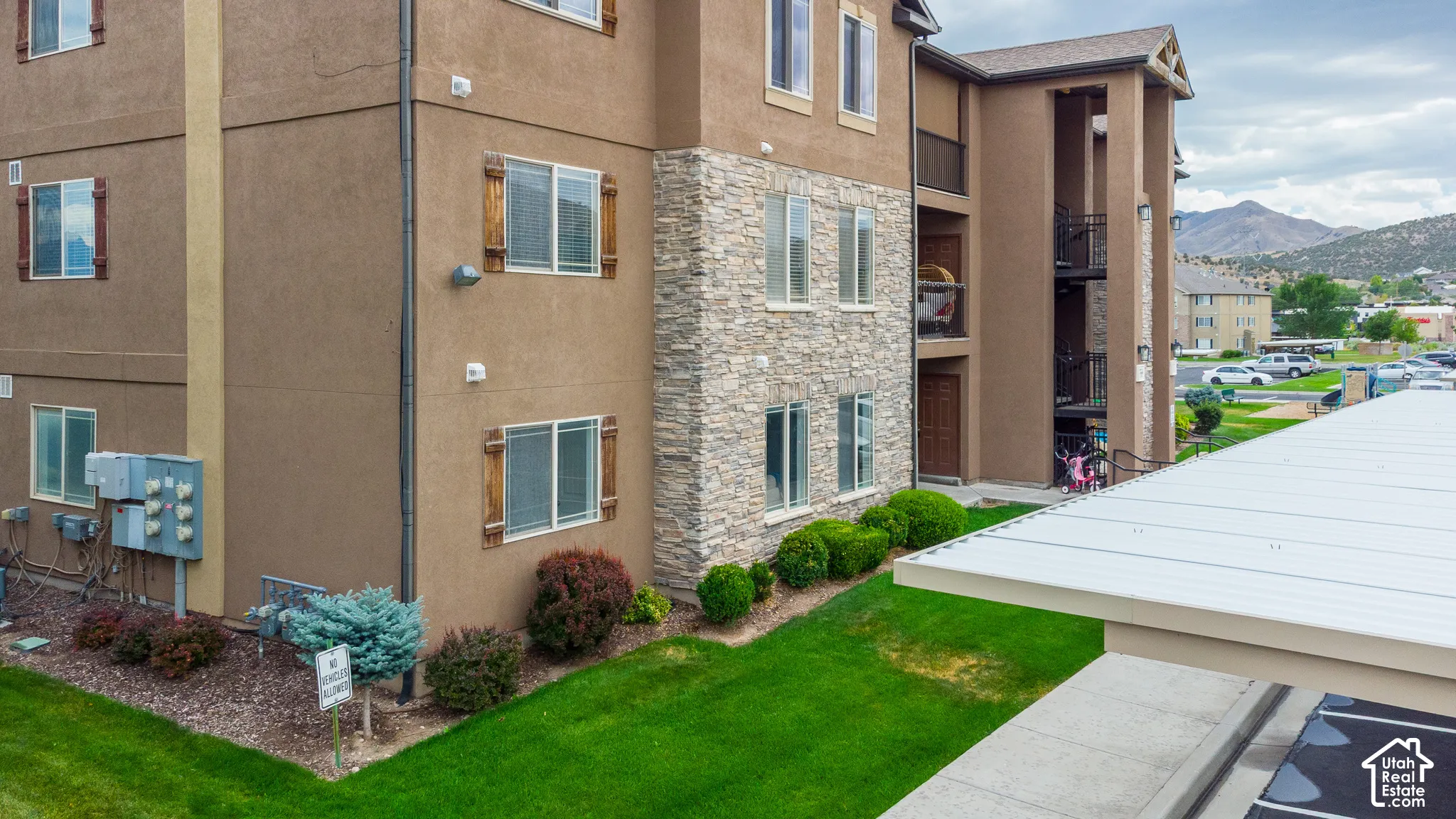 Multi-story exterior featuring stucco siding, stone veneer accents, wood-finish window shutters, a metal-roofed carport, and a mountain backdrop