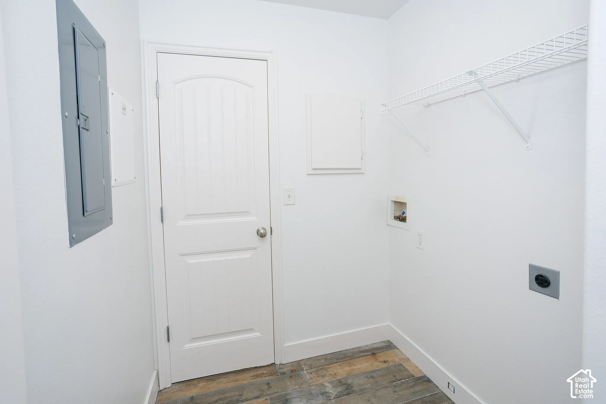 Dedicated utility area featuring wood-finish flooring, a solid-core door, wall-mounted wire shelving, and integrated electrical service panel