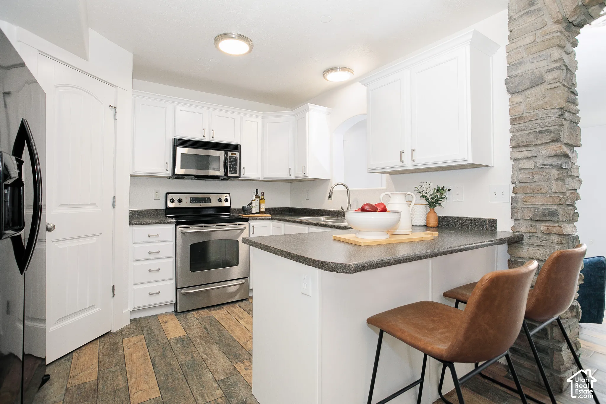 Kitchen featuring white shaker-style cabinetry, stainless steel appliances, dark stone-finish countertops, wood-finish flooring, and a stone-clad arched pillar