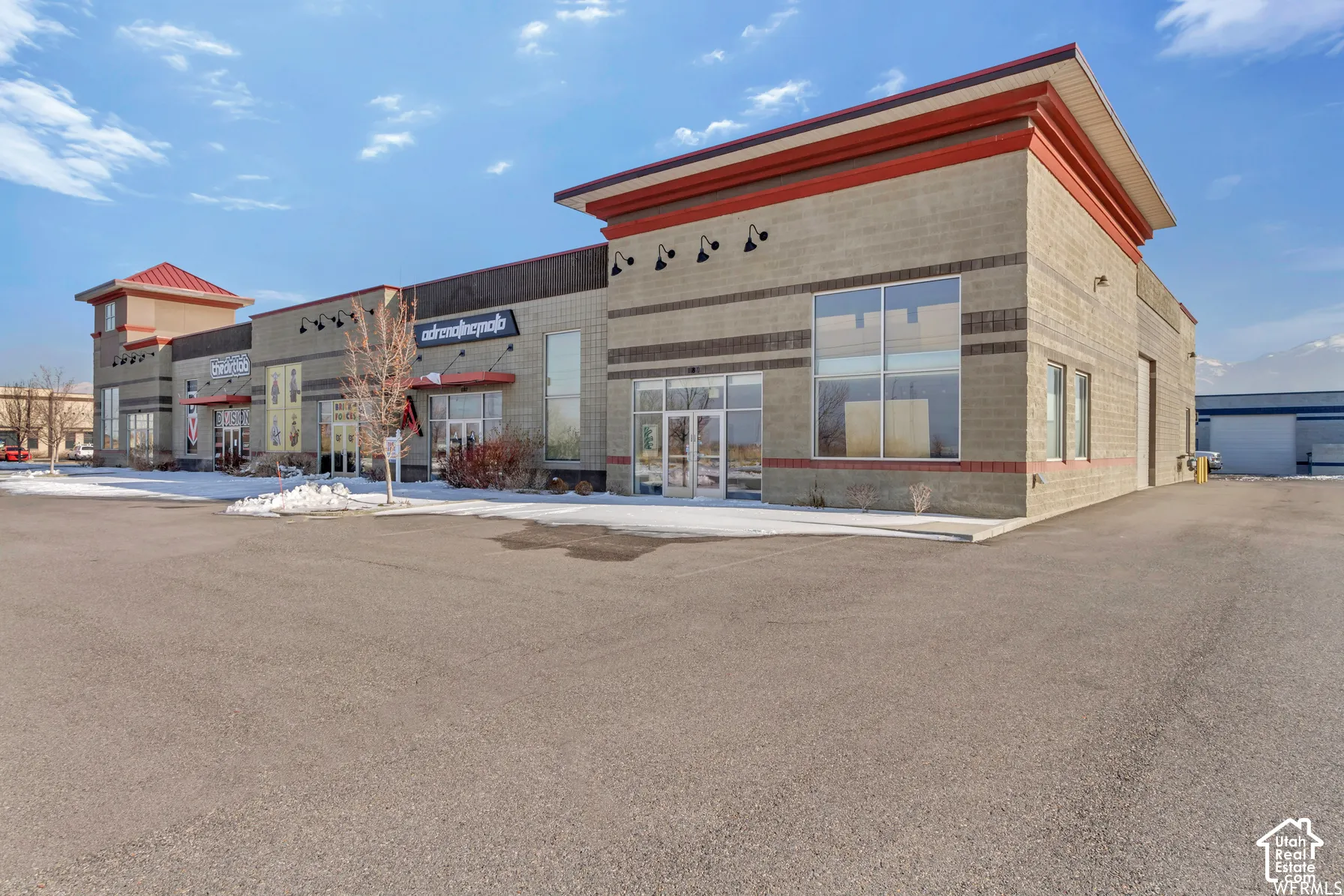 Commercial exterior featuring multi-tenant storefronts, decorative masonry facade, expansive glass entryways, integrated gooseneck lighting, and a paved asphalt parking area