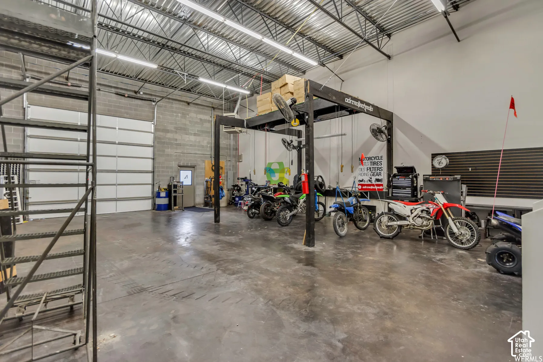 Industrial workspace featuring high-clearance ceilings, exposed metal trussing, concrete block walls, a steel staircase, and overhead garage door access