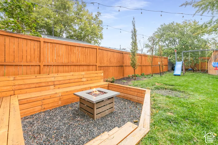 Backyard featuring a wood-slat privacy fence, built-in tiered wood bench seating, a stone-surround fire pit, crushed rock ground cover, and overhead string lighting