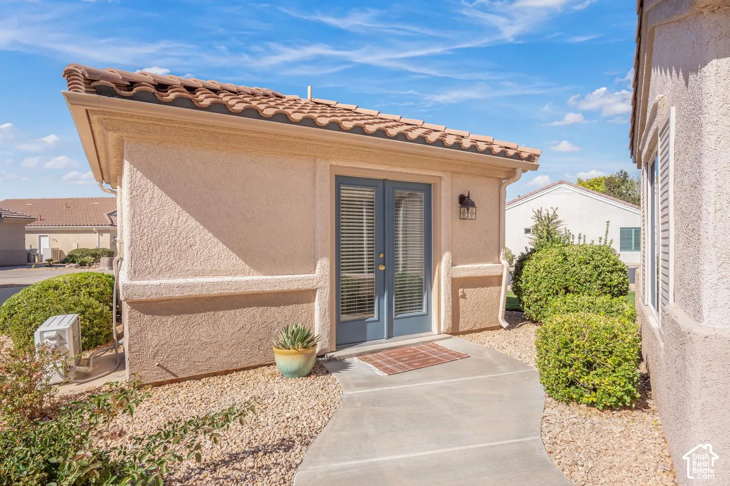 Stucco exterior featuring a clay tile roof, glass-paneled double doors, concrete walkway, and wall-mounted exterior lighting