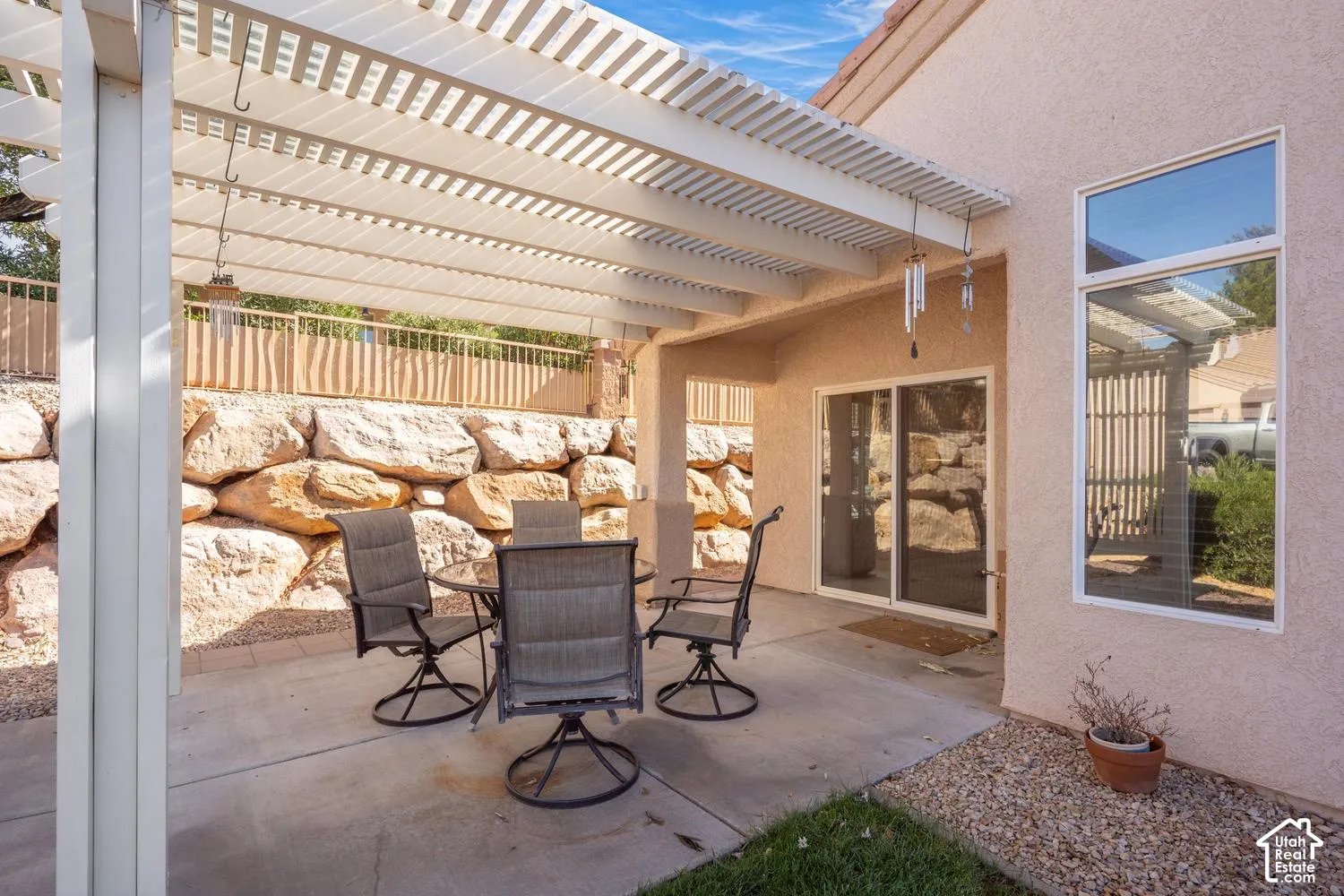 Covered concrete patio featuring a white slatted pergola, decorative stone retaining wall, stucco exterior, and sliding glass door