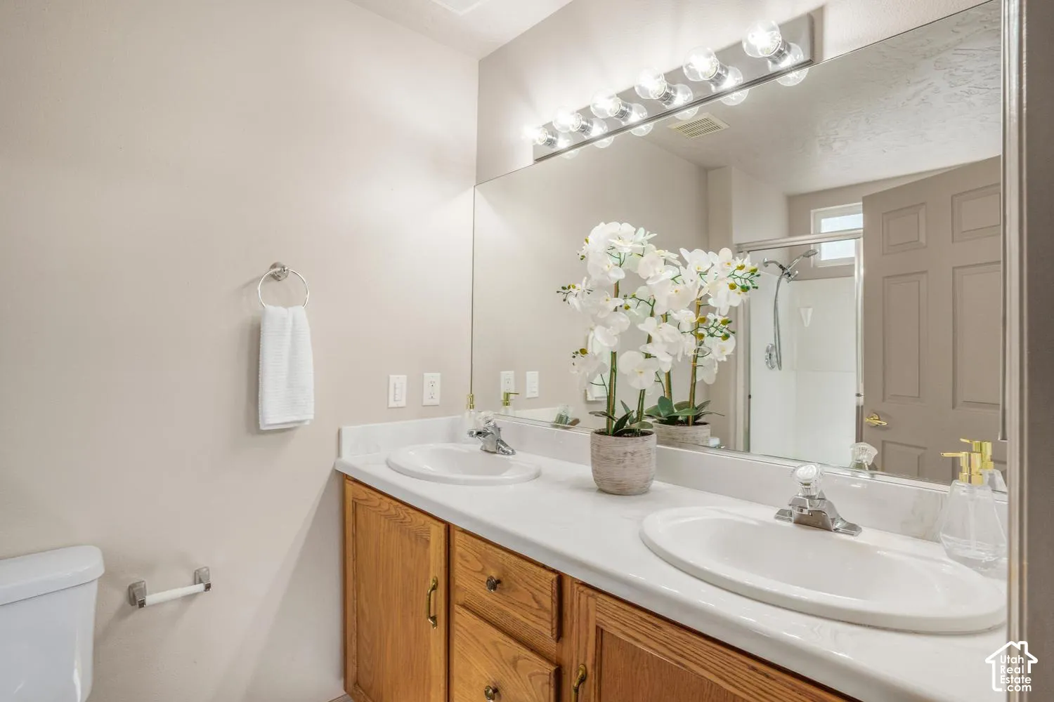 Bathroom featuring a dual-sink vanity with a solid-surface countertop, cabinetry with natural wood finish, a full-width wall mirror, and an overhead multi-bulb light fixture