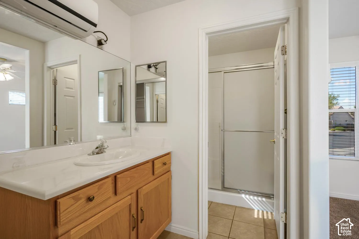 Bathroom featuring a wooden vanity with a white countertop and oval undermount sink, large wall mirror, wall-mounted air conditioning unit, and a tiled shower enclosure with sliding glass doors