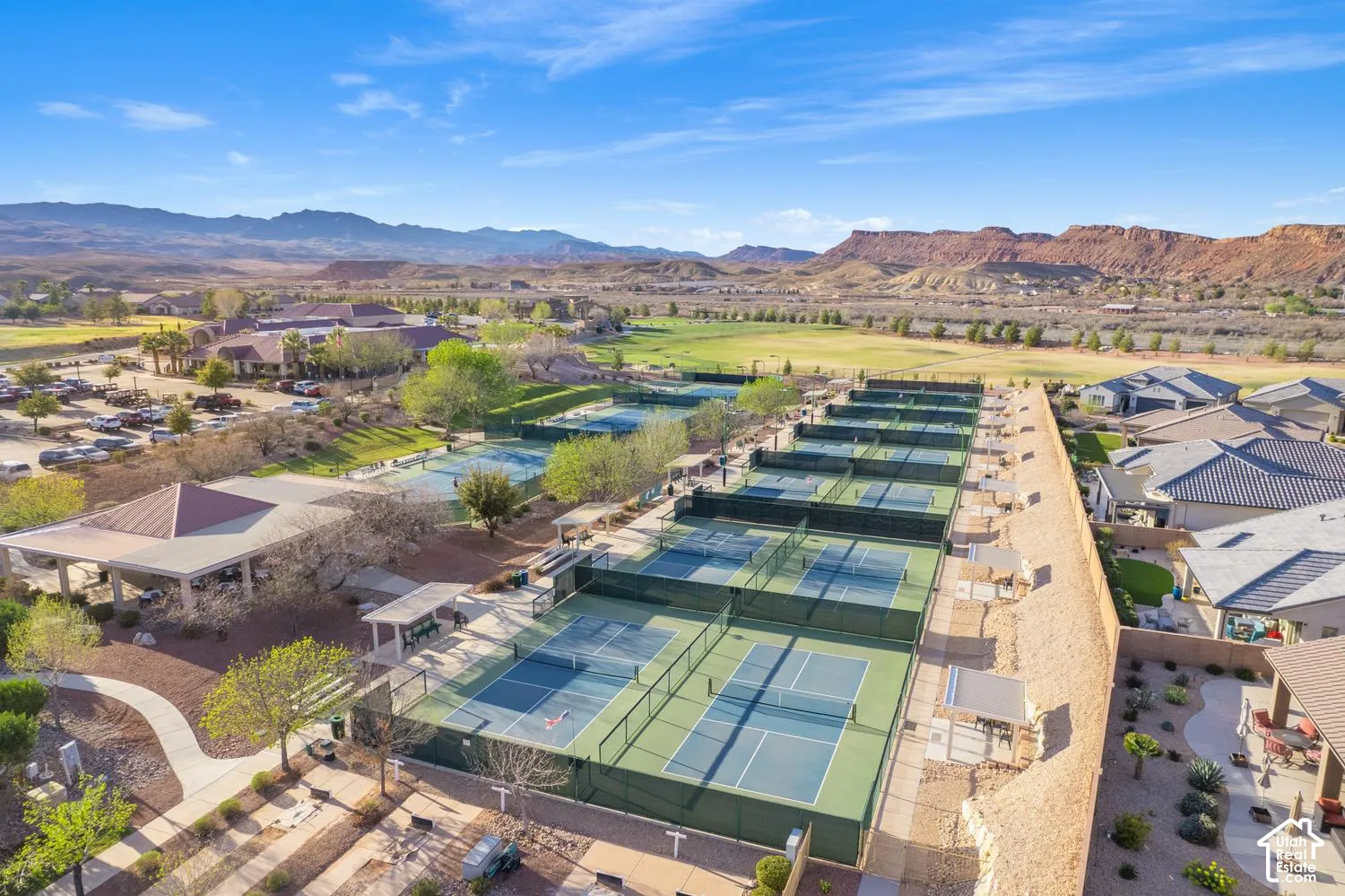 Multiple hard-court tennis and pickleball courts with perimeter fencing, shaded spectator pavilions, concrete walkways, and mountain-range scenery