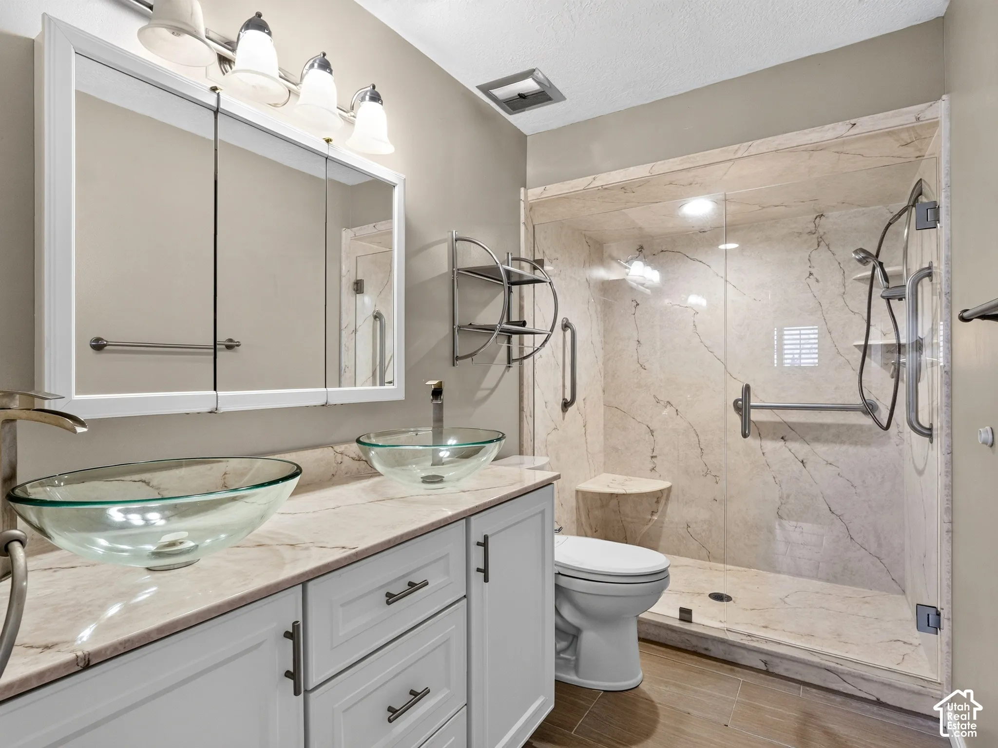 Bathroom featuring white shaker-style cabinetry, dual glass vessel sinks with stone-finish countertops, and a walk-in shower with integrated bench seating and stone-style wall surround