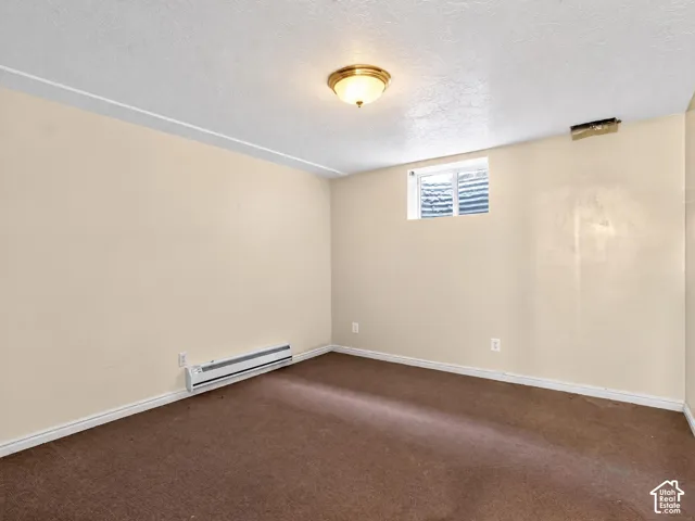 Neutral-toned interior space featuring brown carpeting, a flush-mount ceiling light, a built-in electric baseboard heater, and a rectangular egress window with a window well