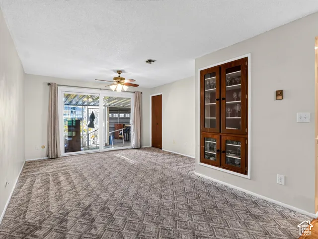 Living area featuring textured carpeting, built-in wooden cabinetry with glass-paneled doors, a ceiling fan with integrated lighting, and a sliding glass door