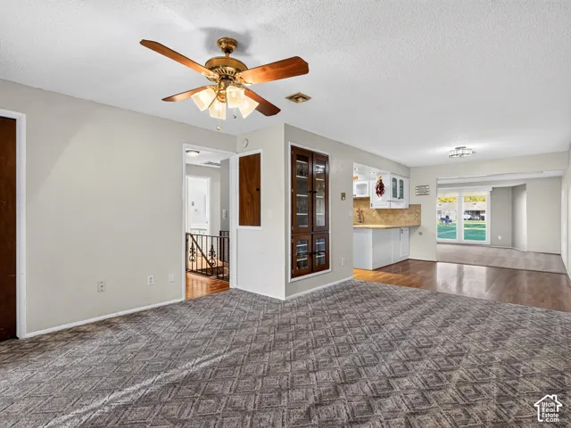 Living space featuring patterned wall-to-wall carpeting, wood-finish flooring in the kitchen, built-in display cabinetry, and a ceiling fan with integrated lighting