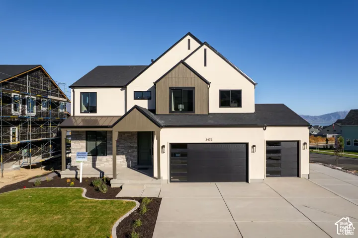 Two-story exterior featuring a light-toned stucco facade, stone veneer accents, wood-finish vertical siding, a metal-accented entry roof, and a three-car garage with glass-paneled doors