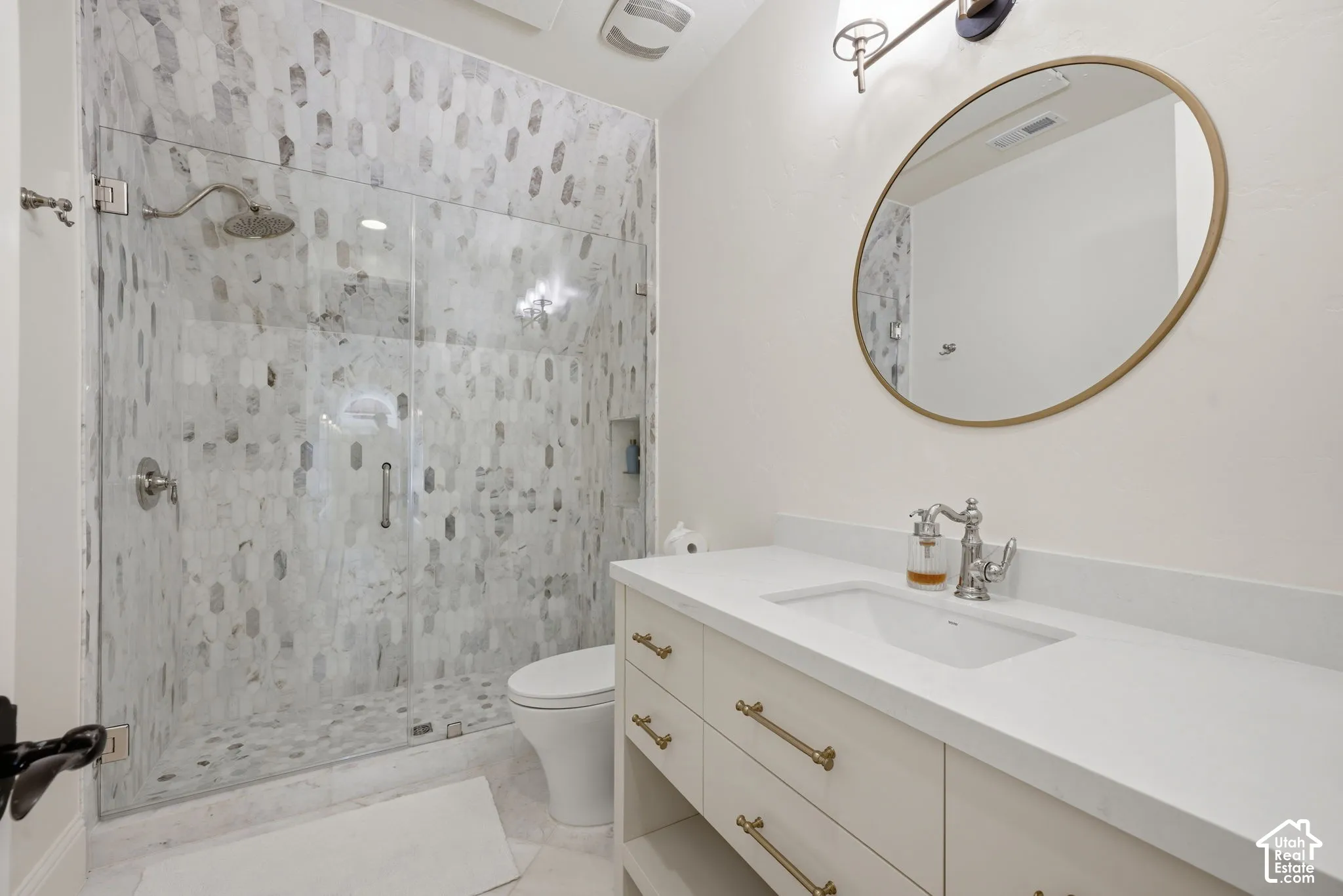 Walk-in shower with patterned tile surround and glass enclosure, light-toned cabinetry, solid surface countertop, integrated vanity sink, and oval wall-mounted mirror