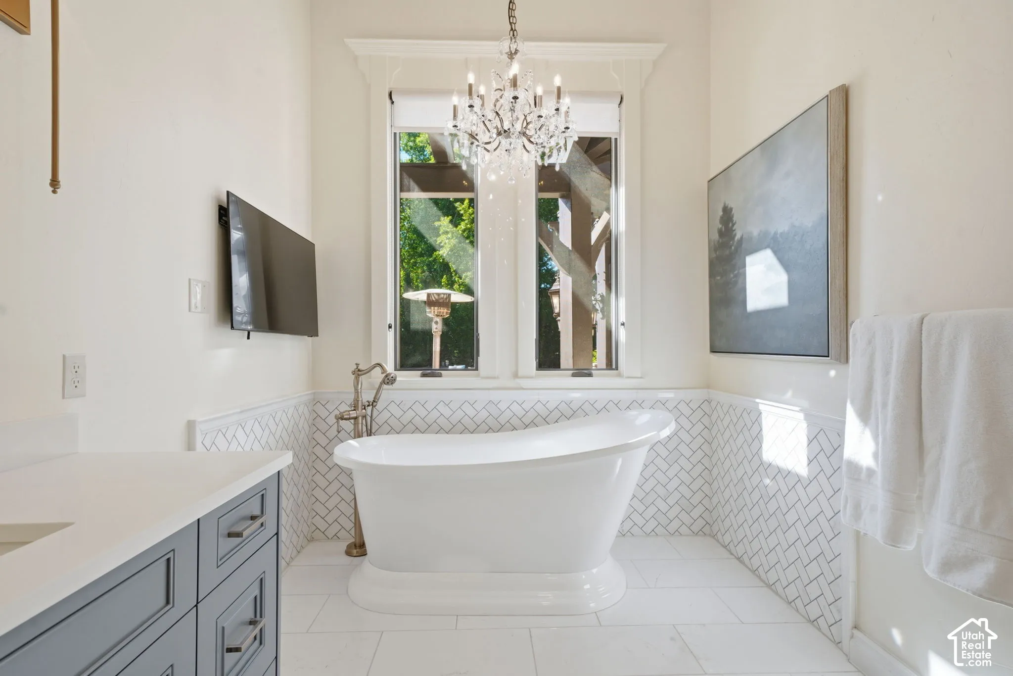 Freestanding soaking tub with floor-mounted faucet, herringbone-patterned wall tile, crystal chandelier, and gray custom vanity with white countertop