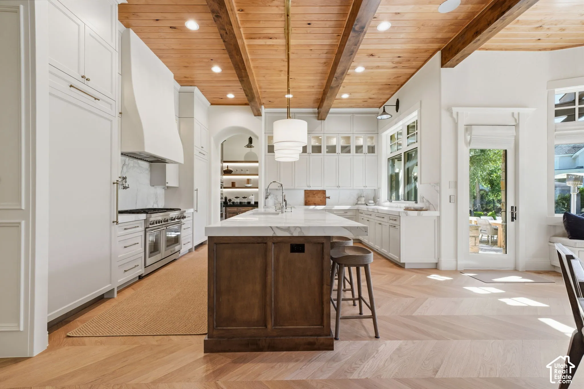 Kitchen featuring a wood-plank ceiling with exposed structural beams, chevron-patterned wood flooring, a central island with a stone countertop, custom cabinetry with integrated paneling, and a gas range with a custom hood