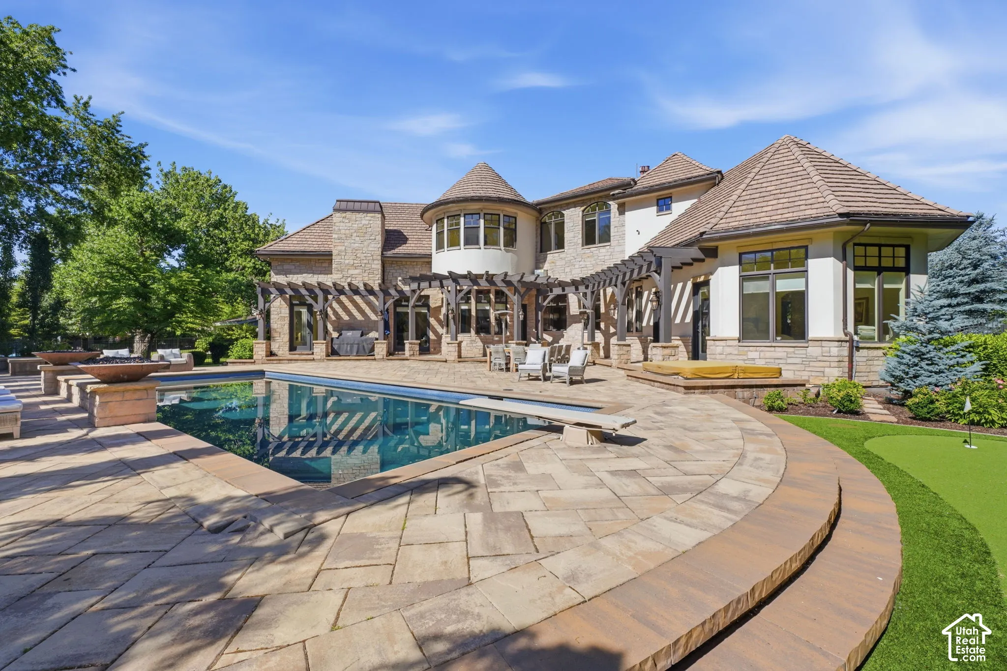 Extensive stone patio featuring an in-ground swimming pool, custom pergola, integrated fire bowls, and a synthetic turf putting green