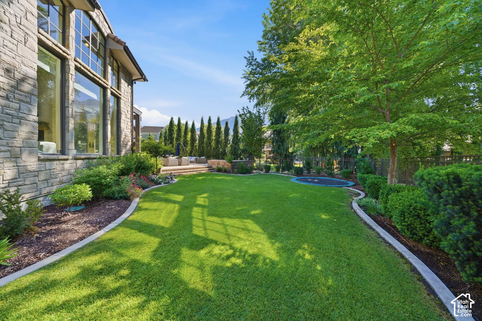 Rear exterior featuring natural stone cladding, oversized windows, manicured lawn, tiered stone patio, and established perimeter landscaping