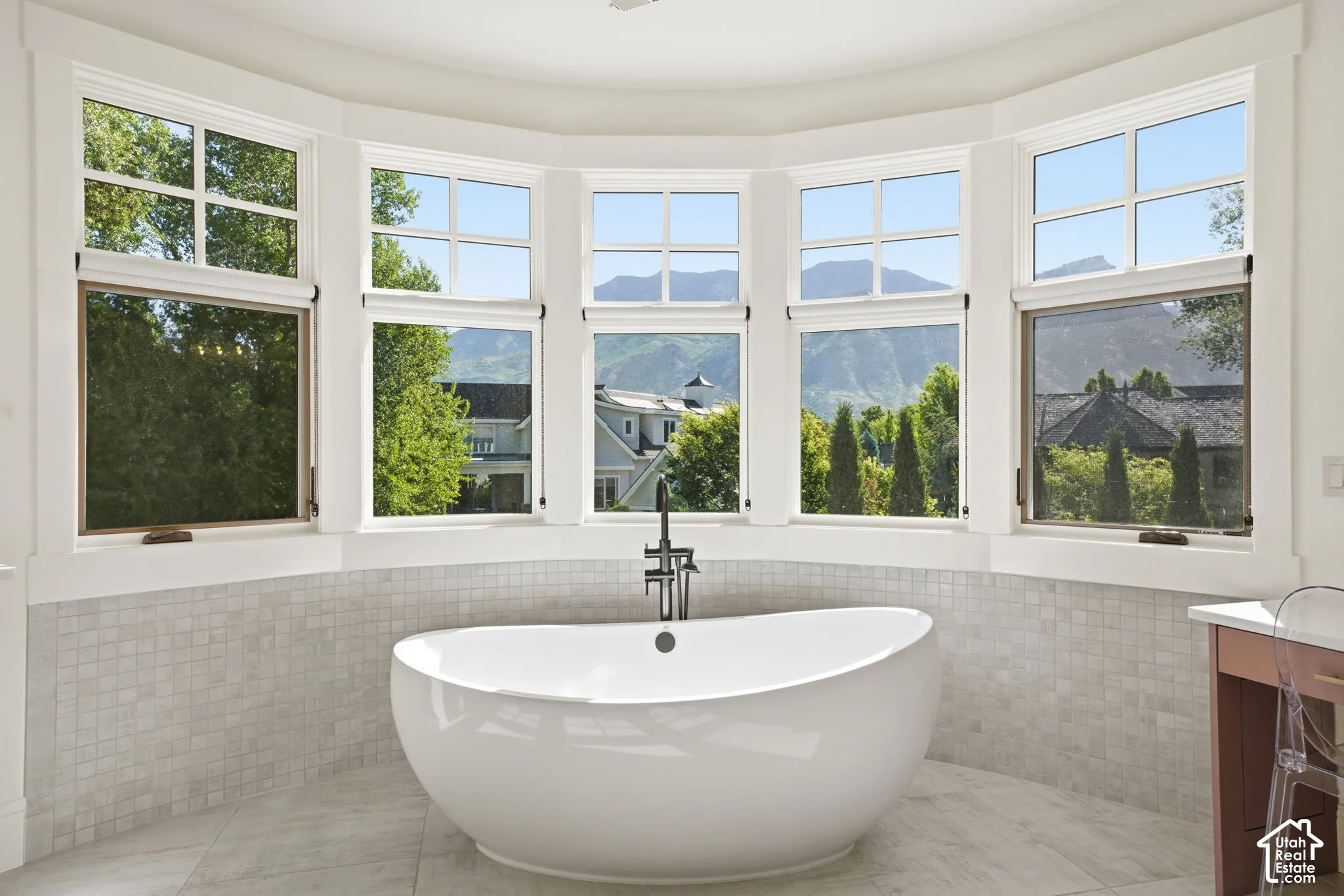 Freestanding white soaking tub with a floor-mounted faucet, set against a multi-panel bay window featuring mountain scenery and stone-tile wainscoting