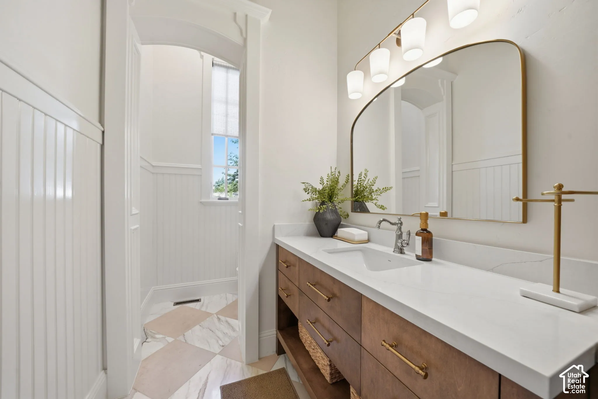 Vanity featuring a light-toned solid surface countertop with an integrated basin, wood-finish cabinetry with metallic hardware, wall-mounted arched mirror, four-light vanity fixture, and vertical beadboard wainscoting