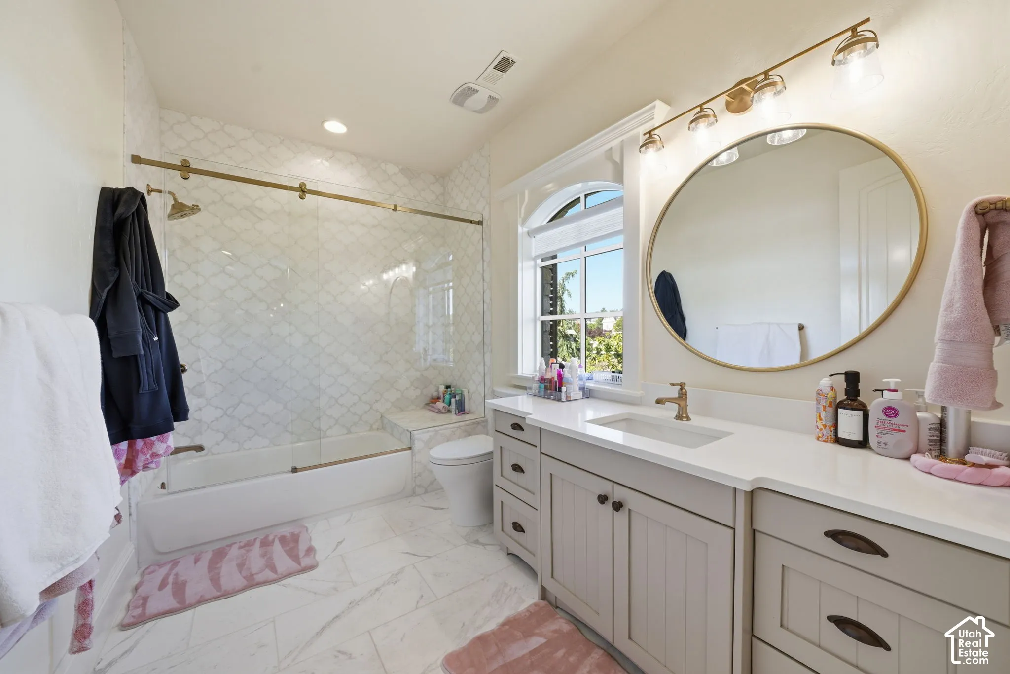 Bathroom featuring a custom vanity with stone-style countertop, gold-tone hardware, arched window, and a shower-tub combination with patterned wall tile and glass enclosure