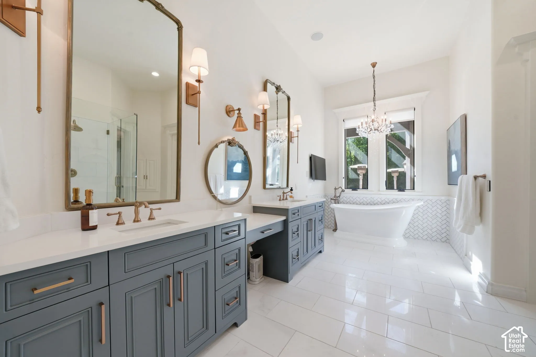 Dual vanities featuring shaker-style cabinetry, quartz countertops, and brass-finish hardware, paired with a freestanding soaking tub, herringbone-patterned tile wall detail, and crystal chandelier