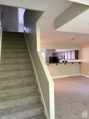 Carpeted staircase with wall-mounted handrail, kitchen featuring wood-finish cabinetry, built-in breakfast bar, and neutral wall color