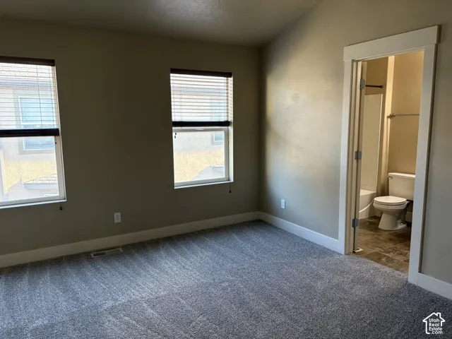 Room featuring vaulted ceilings, carpeted flooring, two windows with horizontal blinds, and a door to an en-suite bathroom with a neutral-tone vanity and tile flooring