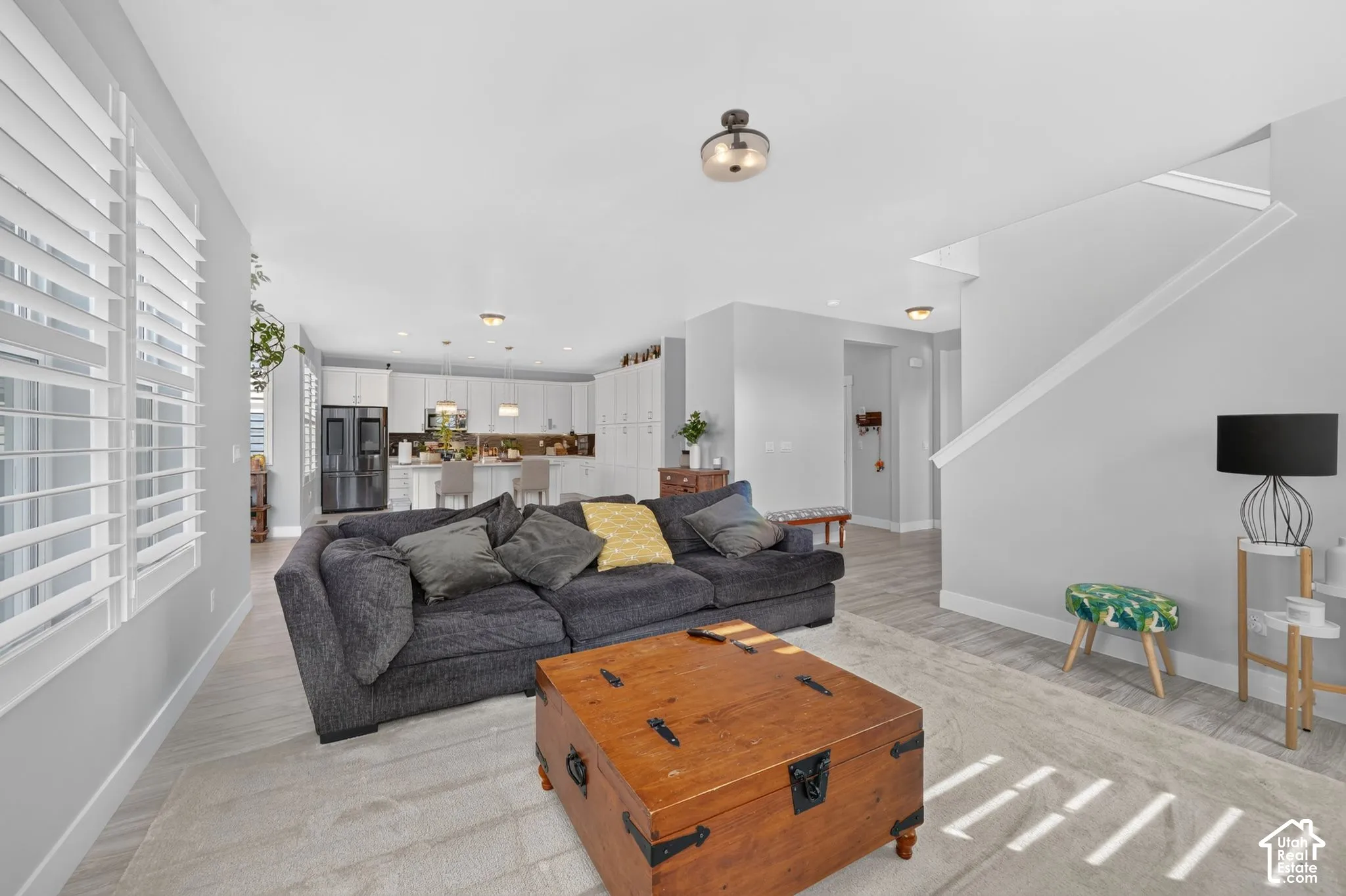 Open-concept interior featuring light wood-finish flooring, white shutter window treatments, a kitchen with white cabinetry, recessed ceiling lighting, and a staircase with a white paneled railing