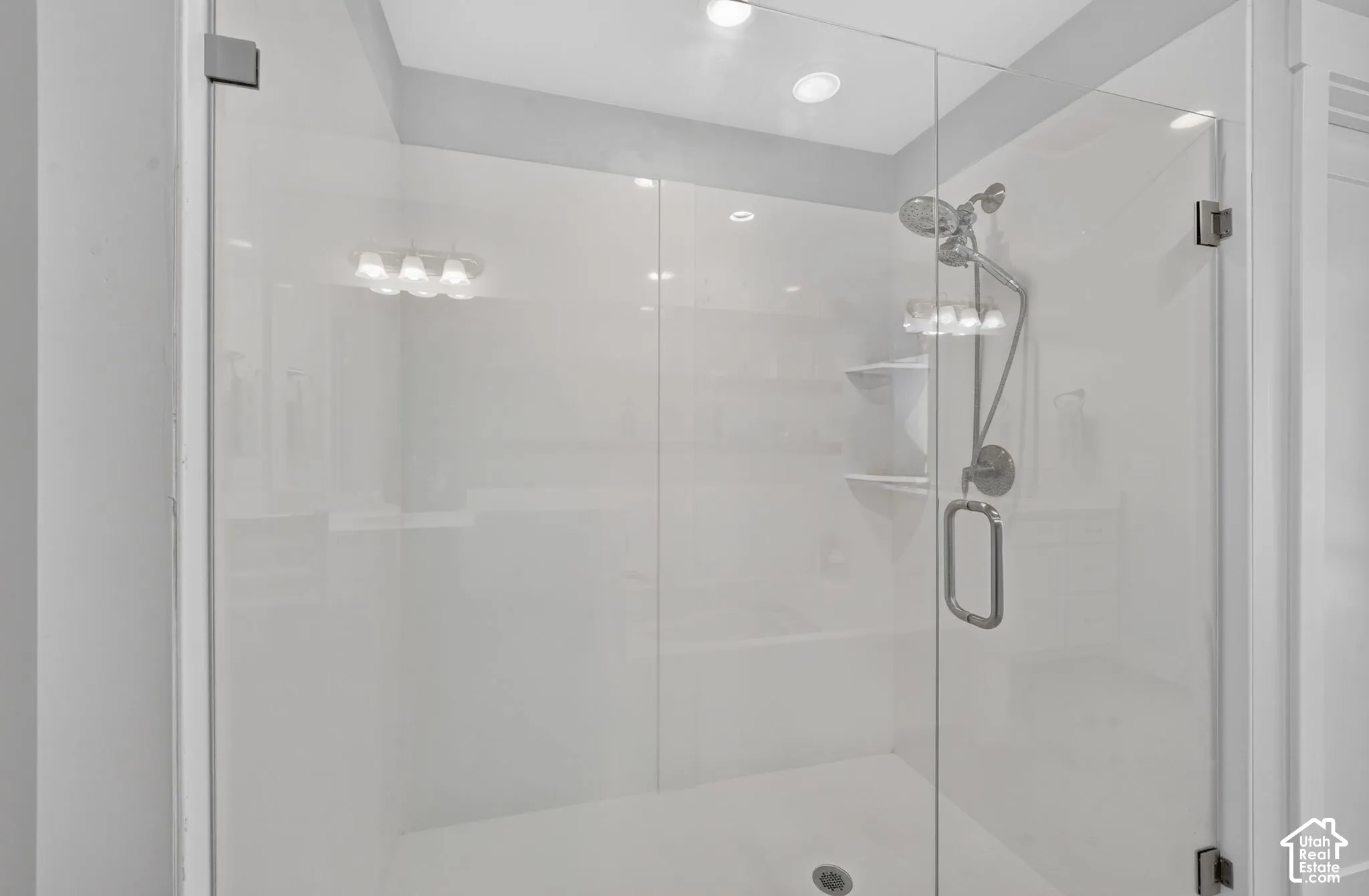 Glass-enclosed shower featuring smooth white wall surrounds, built-in shelving, a dual-head chrome shower fixture, and recessed lighting