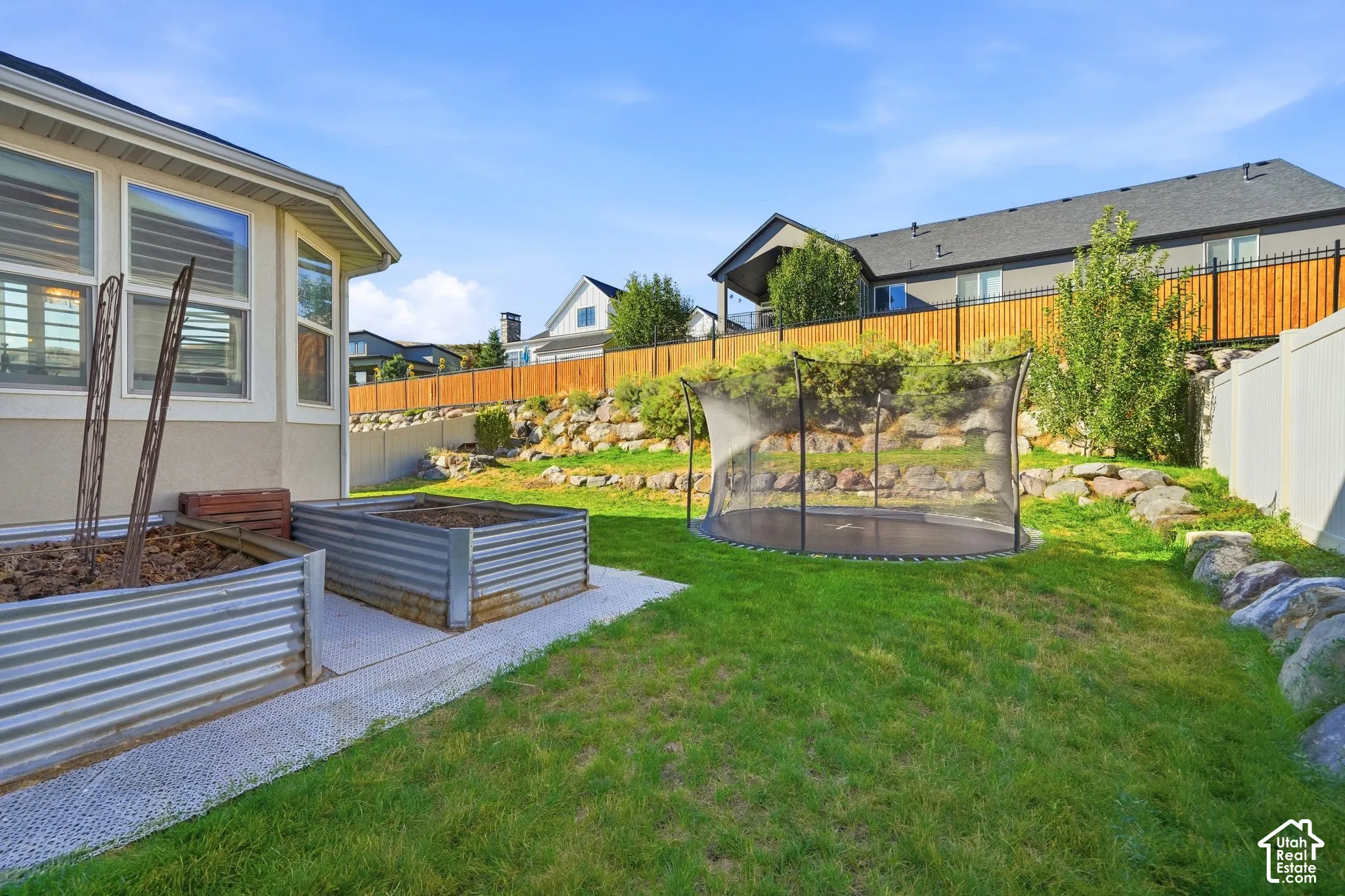 Backyard featuring a tiered stone retaining wall, raised metal garden beds, a gravel-surfaced patio area, and perimeter privacy fencing