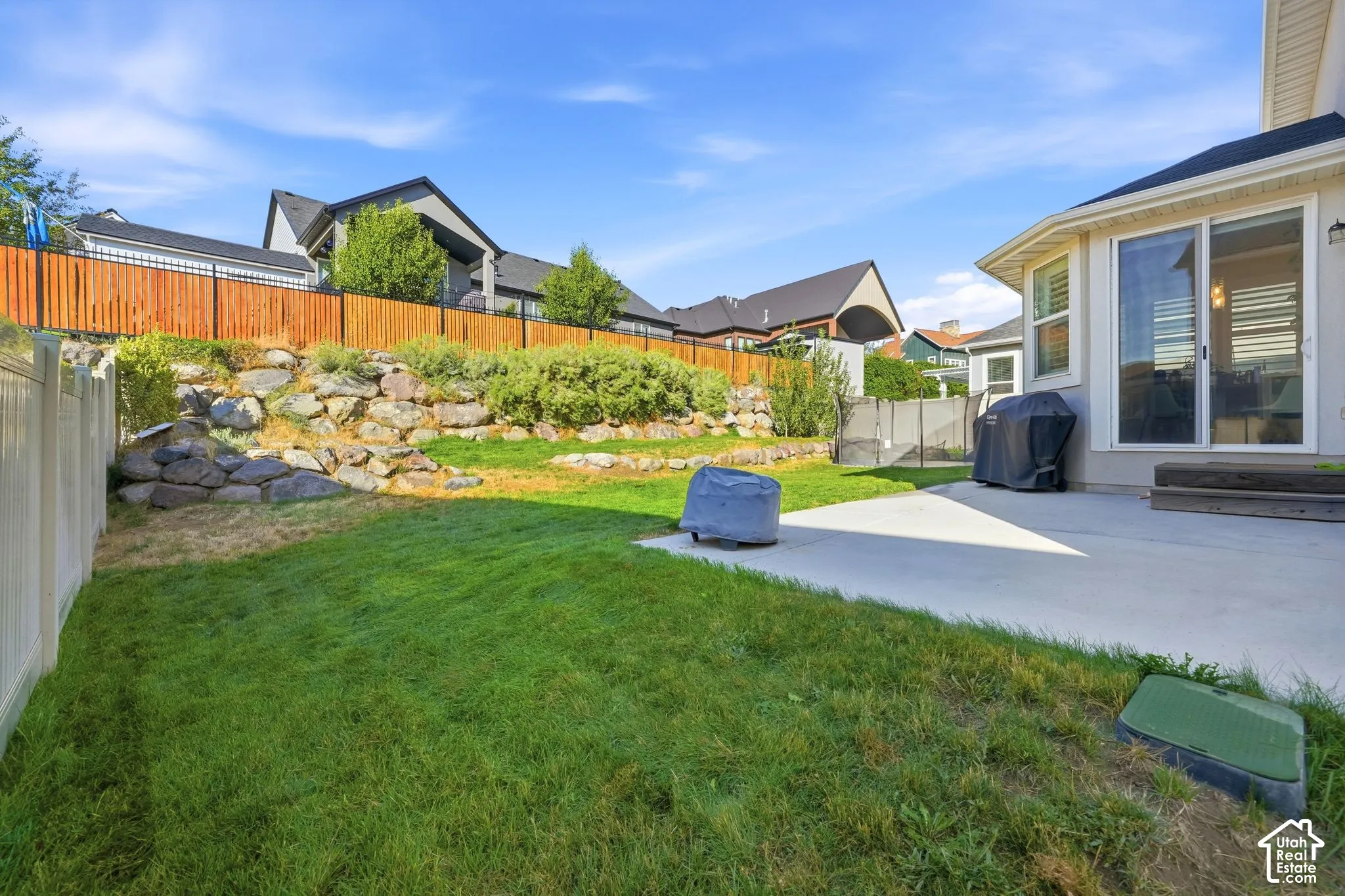 Rear exterior space featuring a concrete patio, tiered stone retaining walls, manicured lawn, and wood-slat perimeter fencing