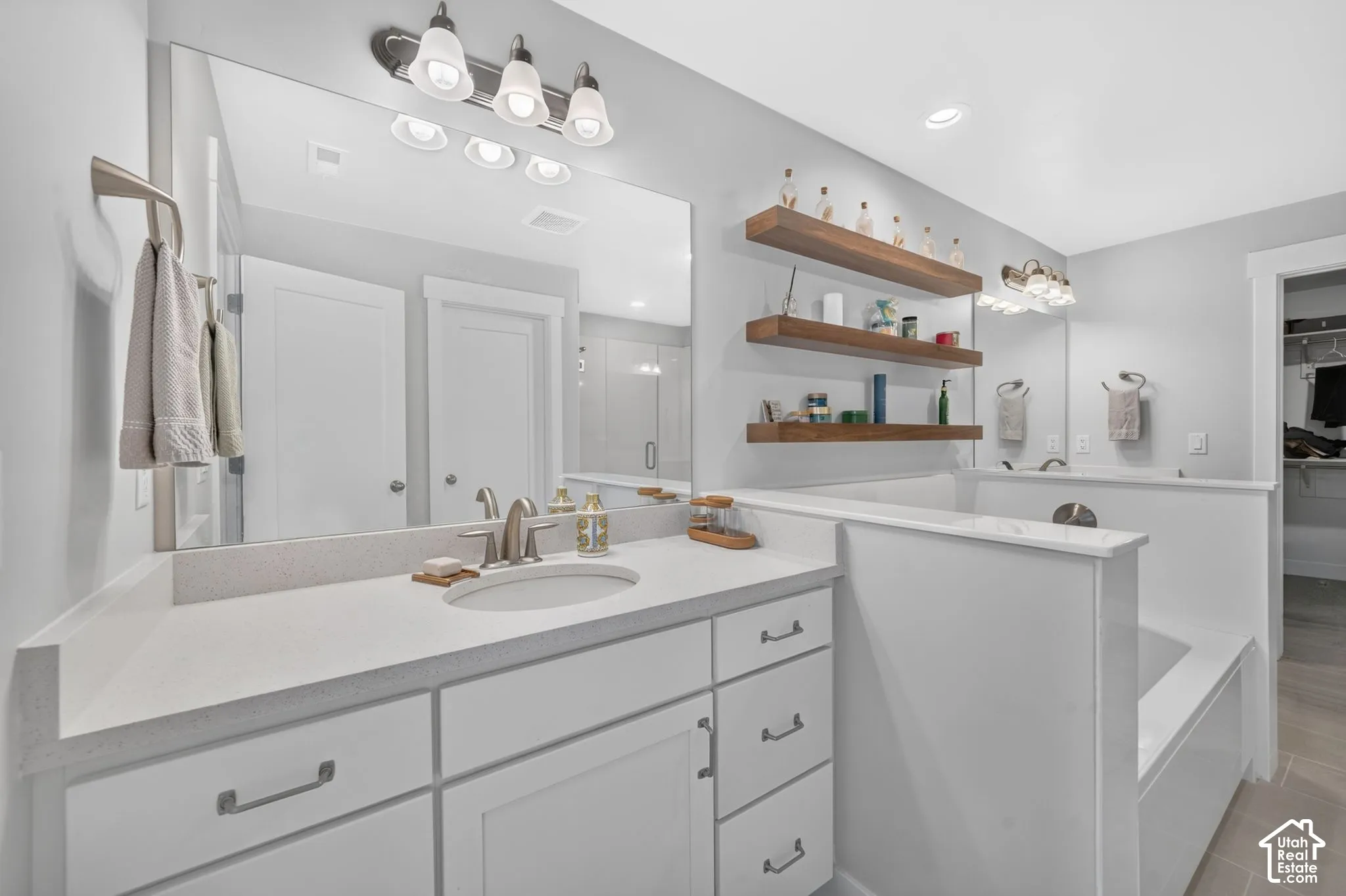 Bathroom featuring a white vanity with composite countertop, undermount sink, large wall-mounted mirror, three-tier wood shelving, and a soaking tub