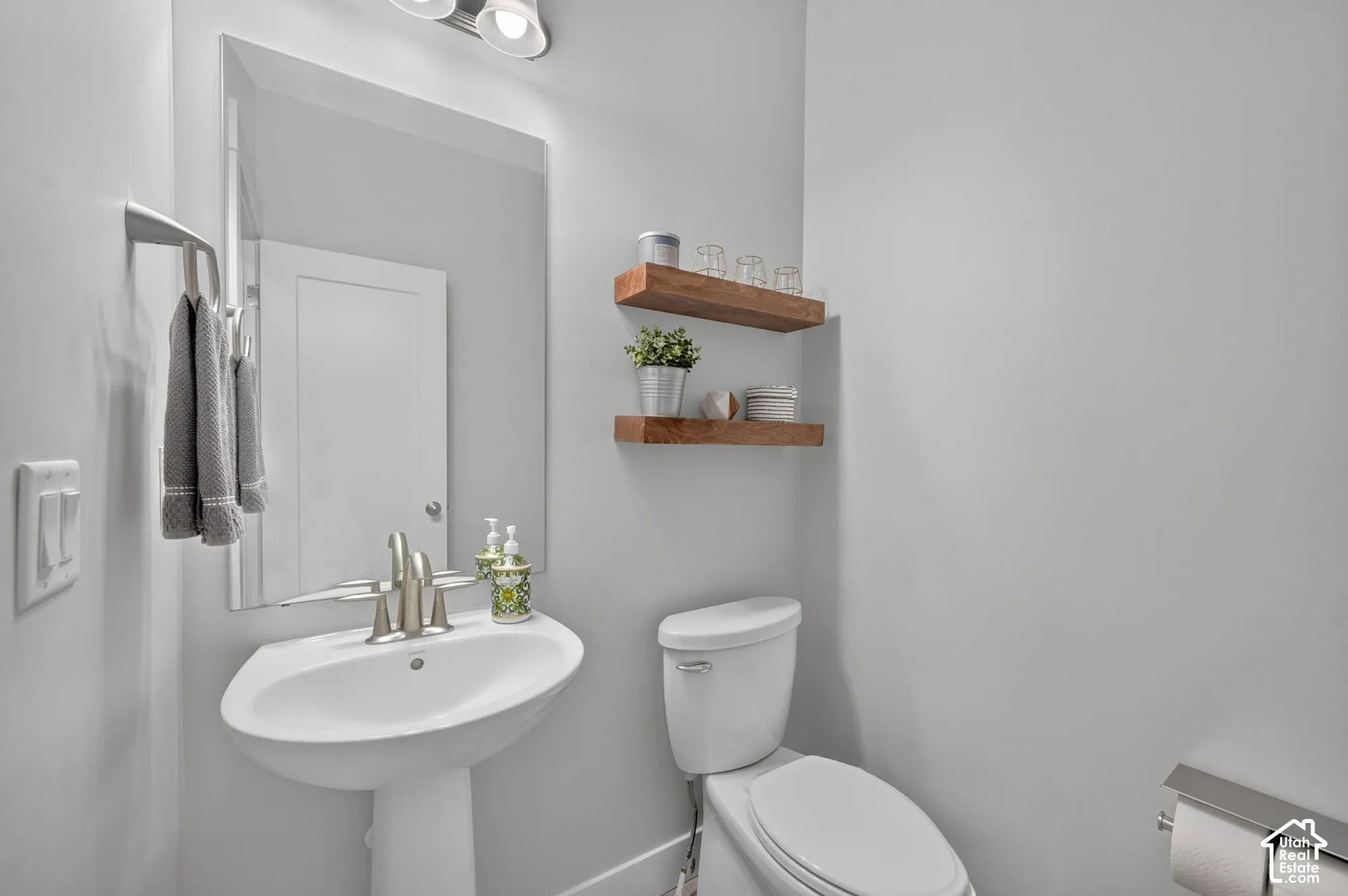 Wall-mounted pedestal sink with polished chrome faucet, expansive mirror, wood-finish shelving, and overhead vanity lighting