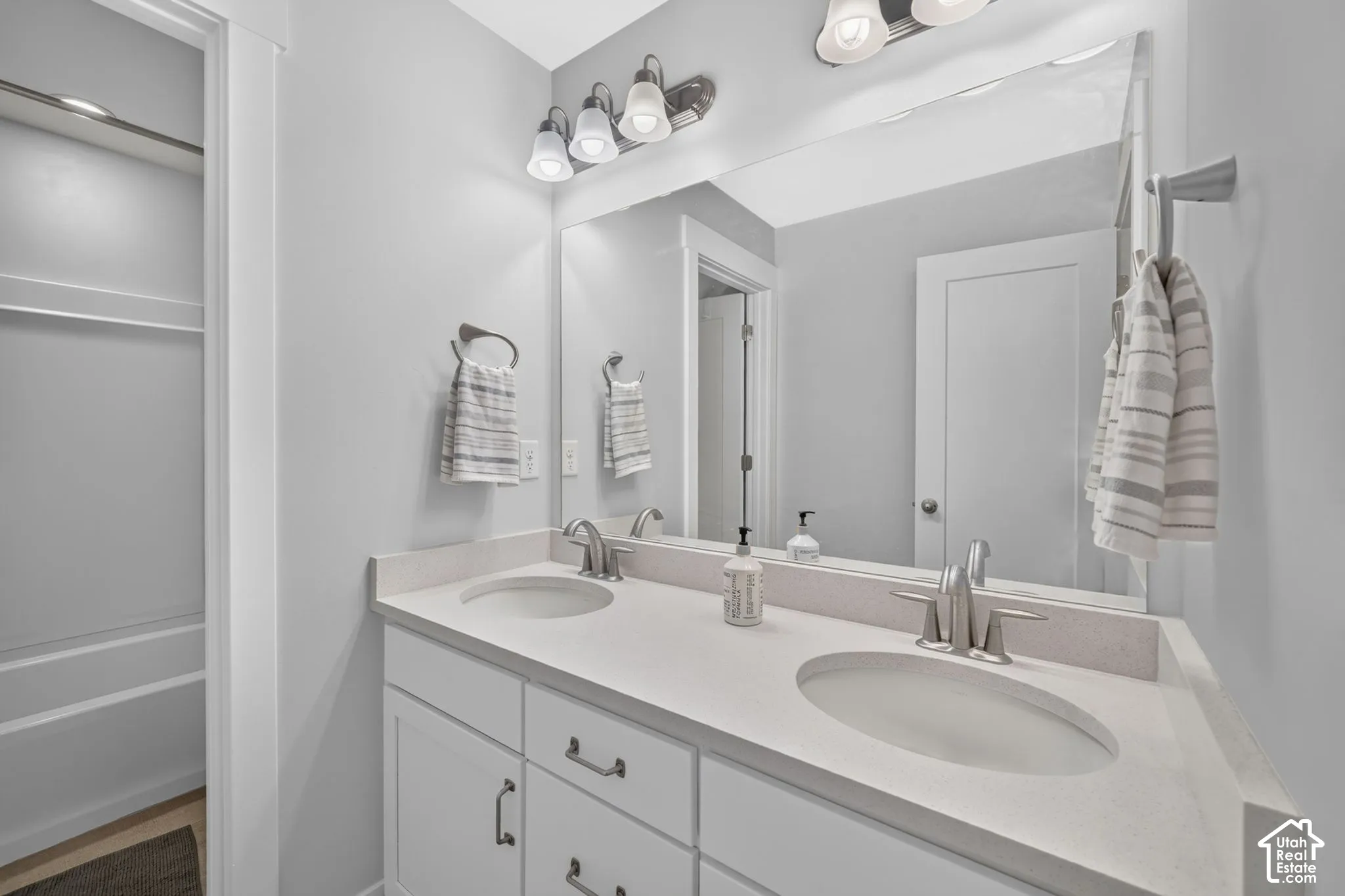 Bathroom vanity featuring dual under-mount sinks, light-toned stone-surface countertop, white shaker-style cabinetry, a full-width mirror, and multi-bulb wall lighting