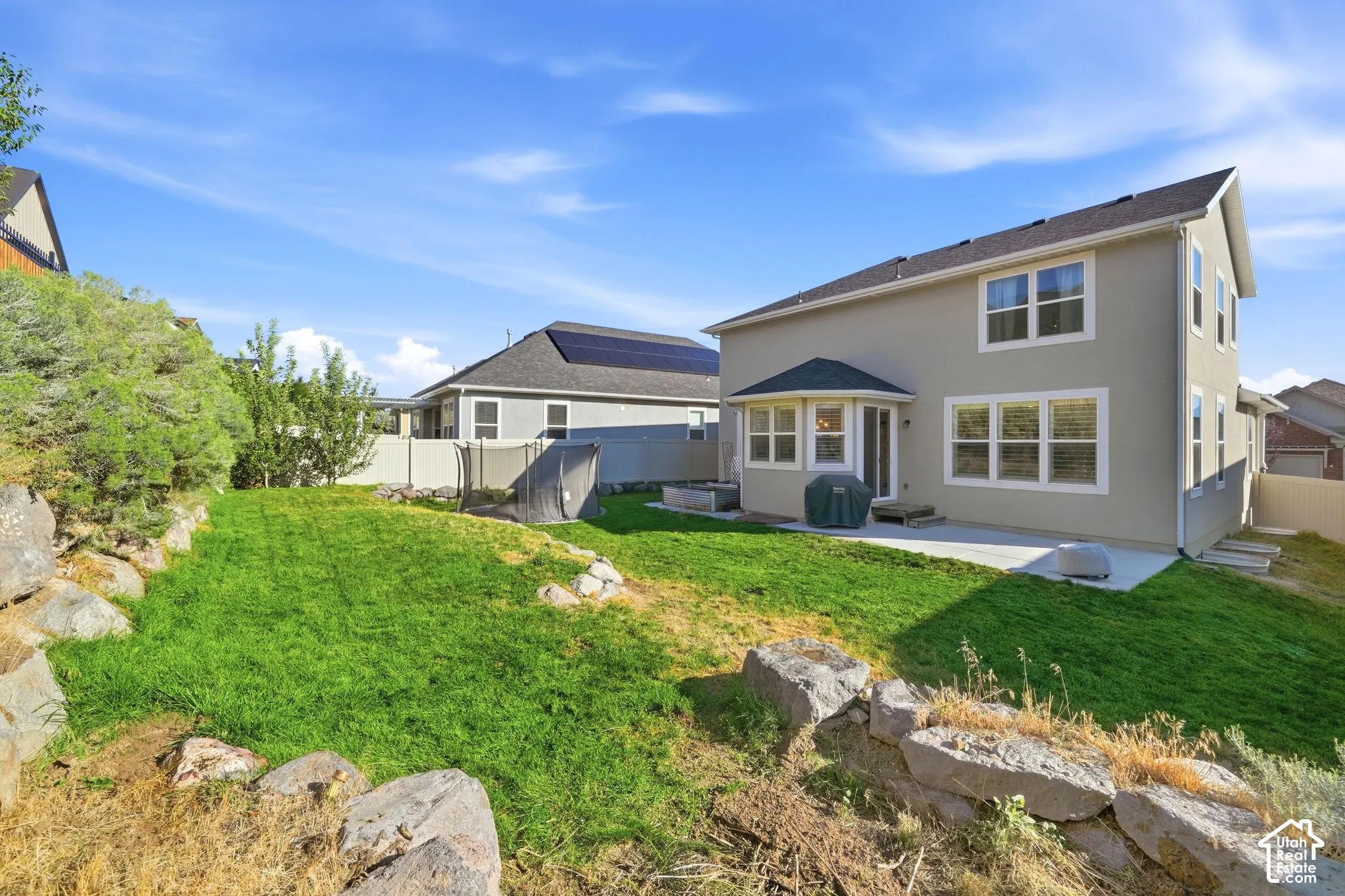 Rear exterior featuring a concrete patio, multi-level sloped landscaping, natural stone retaining walls, and vinyl privacy fencing