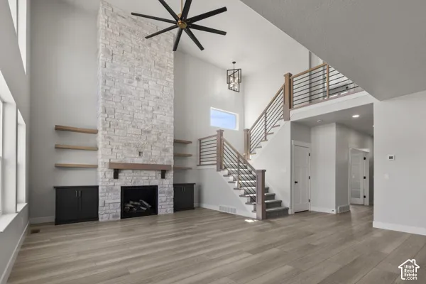 High-ceilinged room featuring a floor-to-ceiling stone fireplace, wood-finish flooring, built-in cabinetry with floating shelving, and an open staircase with horizontal metal railing