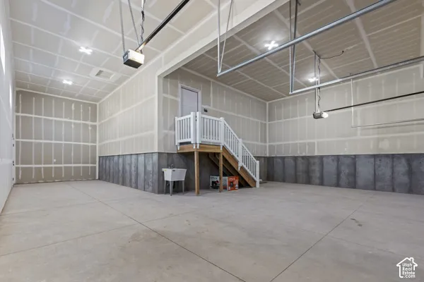 Spacious multi-vehicle garage featuring concrete flooring, finished drywall, overhead lighting, an integrated utility sink, and a staircase with white balusters