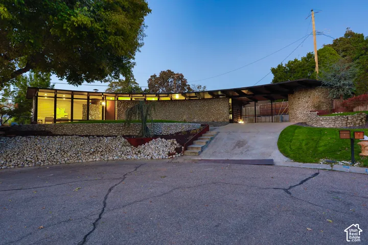 Mid-century modern architectural exterior featuring a curved roofline, floor-to-ceiling glass paneling, extensive natural stone cladding, and a built-in covered carport