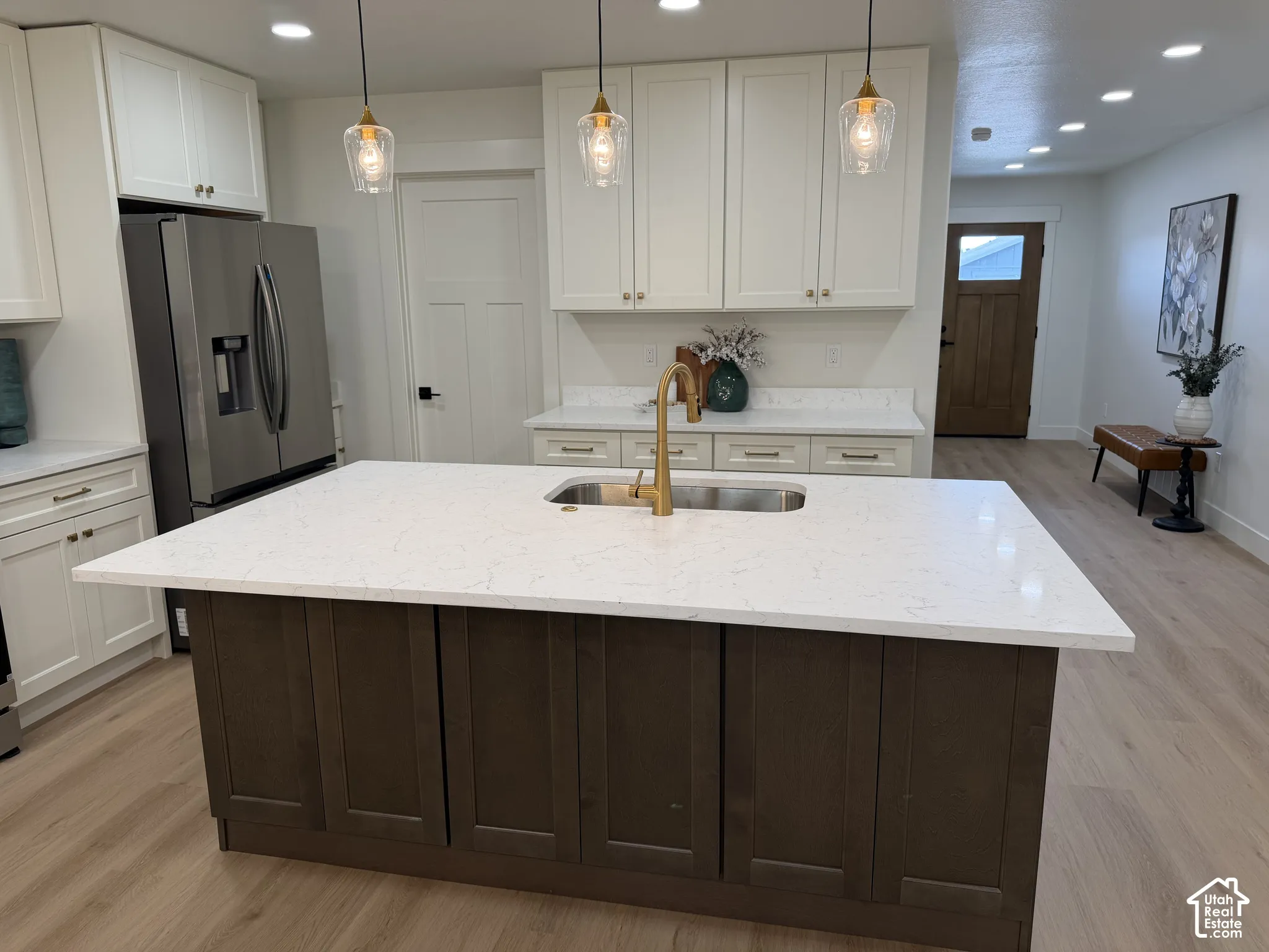 Kitchen featuring an island with a light-toned solid surface countertop and undermount sink, brass-finish faucet, recessed lighting, white cabinetry, and wood-finish flooring