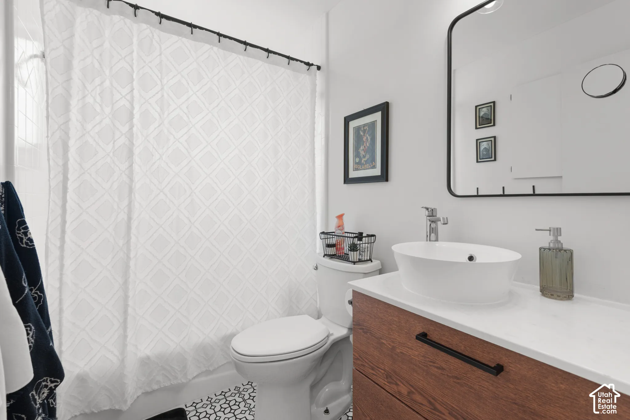 Bathroom featuring a wood-finish vanity with a white vessel sink, quartz-style countertop, rectangular mirror with a black frame, and patterned tile flooring