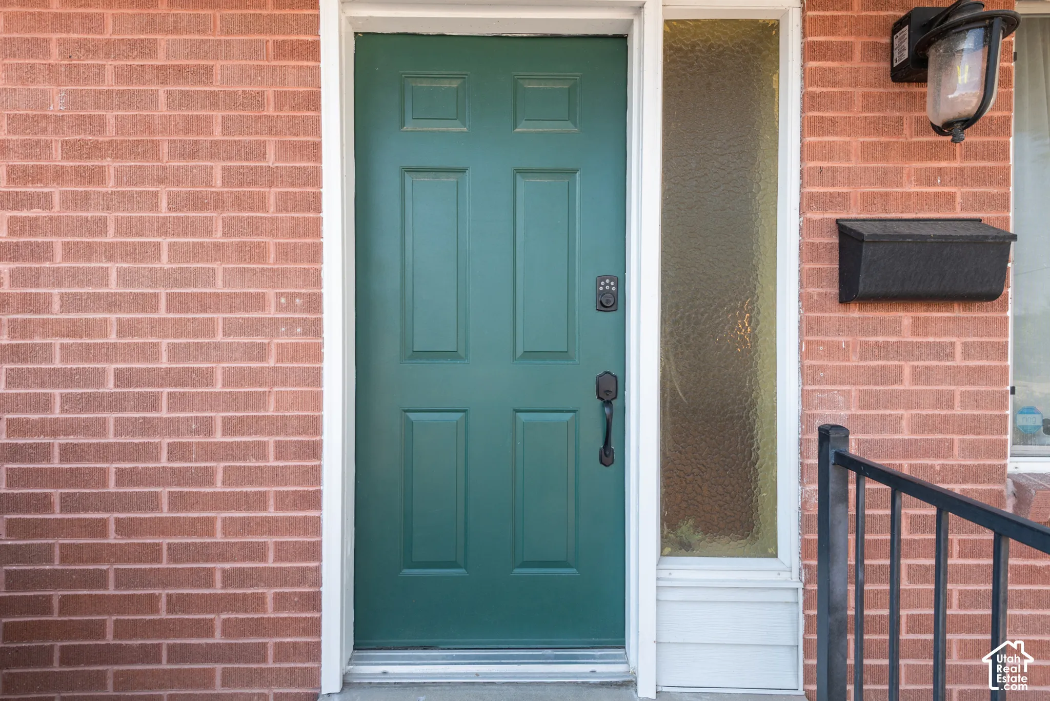 Exterior entrance featuring a six-panel green door, vertical textured glass sidelight, red brick masonry, matte black hardware with electronic deadbolt, and an attached wall-mounted lantern